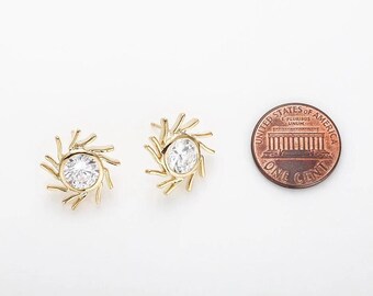 May include: Two gold-colored stud earrings with a sunburst design, each featuring a clear, round gemstone center. A US penny is next to the earrings for size comparison. The earrings are on a white background.