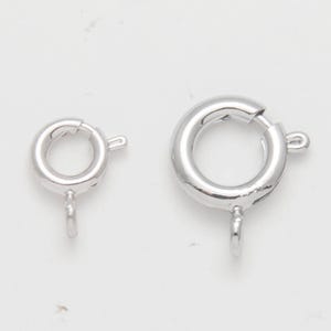 May include: Two silver-colored spring ring clasps of different sizes are displayed against a white background. The clasps are round with a small lever to open and close, and a loop for attaching to jewelry.