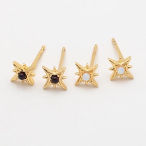 May include: Four gold-colored starburst stud earrings. Two earrings have a small, dark red gemstone in the center, and the other two have a white gemstone. The earrings are on a white background.