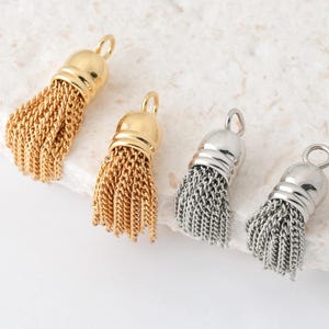 Könnte beinhalten: Vier Metallquastenanhänger in Gold und Silber. Die Anhänger haben eine Schlaufe zum Aufhängen und eine Kettenfransen. Zwei sind goldfarben und zwei silberfarben, alle mit einer glatten, abgerundeten Kappe.