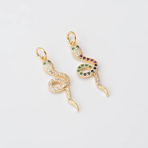 May include: Two gold-colored snake pendants. One snake is covered in clear crystals, the other has a rainbow of colored gems. Both have a loop at the top for hanging. The pendants are on a white background.