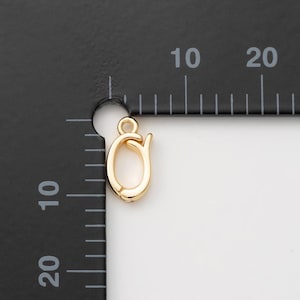 Puede incluir: Un cierre de joyería dorado, con forma de letra 'O' estilizada con un lazo en la parte superior para la fijación. El cierre se muestra sobre un fondo negro con marcas de medición.