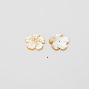 Puede incluir: Dos cuentas de n&aacute;car con forma de flor. Cada cuenta tiene cinco p&eacute;talos de color blanco nacarado y un sutil borde amarillo-anaranjado. El n&uacute;mero "7" est&aacute; impreso en negro debajo de las cuentas.