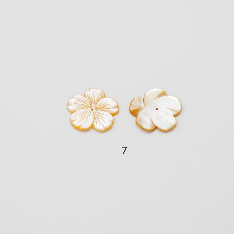 Puede incluir: Dos cuentas de n&aacute;car con forma de flor. Cada cuenta tiene cinco p&eacute;talos de color blanco nacarado y un sutil borde amarillo-anaranjado. El n&uacute;mero "7" est&aacute; impreso en negro debajo de las cuentas.