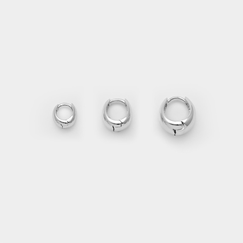 May include: Three silver hoop earrings of different sizes are shown against a white backdrop. The earrings have a smooth, rounded design with a small clasp. The sizes appear to be small, medium, and large.