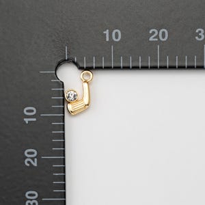May include: A gold-colored golf club charm with a clear, round crystal ball. The charm has a loop at the top for attaching to a necklace or bracelet. The golf club is detailed with horizontal lines.