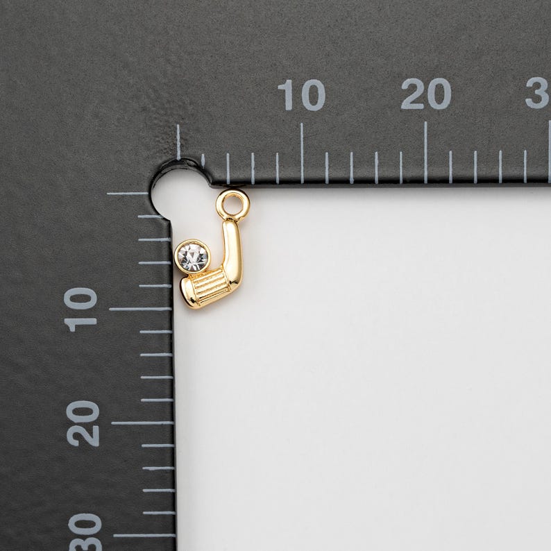May include: A gold-colored golf club charm with a clear, round crystal ball. The charm has a loop at the top for attaching to a necklace or bracelet. The golf club is detailed with horizontal lines.