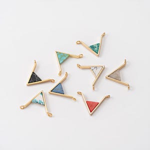 Op de afbeelding: Een verzameling goudkleurige metalen driehoekige bedels met verschillende stenen inlays. De inlays hebben kleuren als turquoise, groen, wit, zwart, blauw en rood. Elke bedel heeft aan elk uiteinde een lus om aan sieraden te bevestigen.