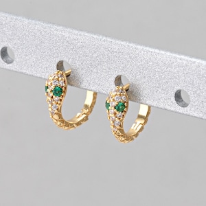 May include: A pair of gold-colored hoop earrings. Each earring features a snake design with a head adorned with green and clear gemstones. The earrings are displayed on a silver metal bar.