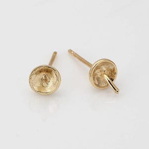 May include: Two gold-colored stud earrings. Each earring features a small, textured, cup-shaped design with a post for insertion. The earrings are set against a white background, highlighting their simple elegance.
