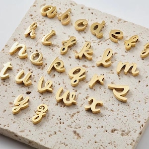 Op de afbeelding: Goudkleurige kleine letter alfabet bedels gerangschikt op een getextureerd, lichtgekleurd oppervlak. De letters zijn in een cursief lettertype, waarbij elke letter duidelijk gedefinieerd is. Ideaal voor het maken van sieraden of gepersonaliseerde knutselwerkjes.