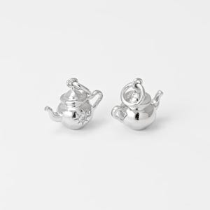 May include: Two small, shiny silver teapot charms. Each charm has a handle, spout, and a lid with a loop for attaching to a chain or bracelet. One teapot has a starburst design on its side. The charms are on a white background.