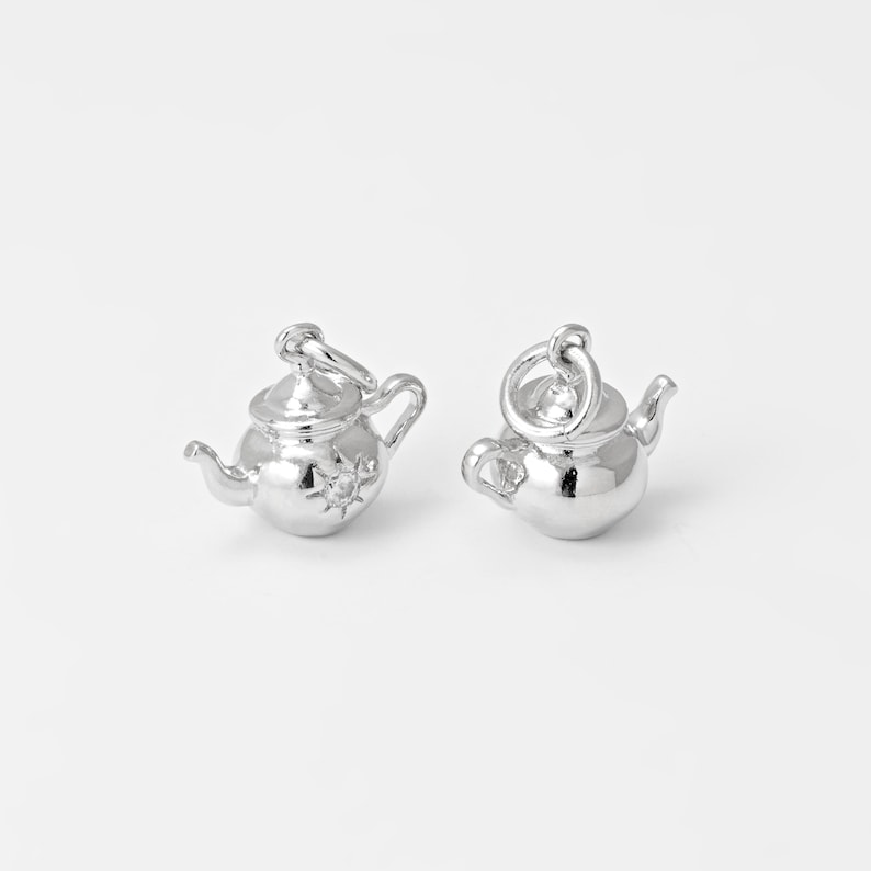 May include: Two small, shiny silver teapot charms. Each charm has a handle, spout, and a lid with a loop for attaching to a chain or bracelet. One teapot has a starburst design on its side. The charms are on a white background.