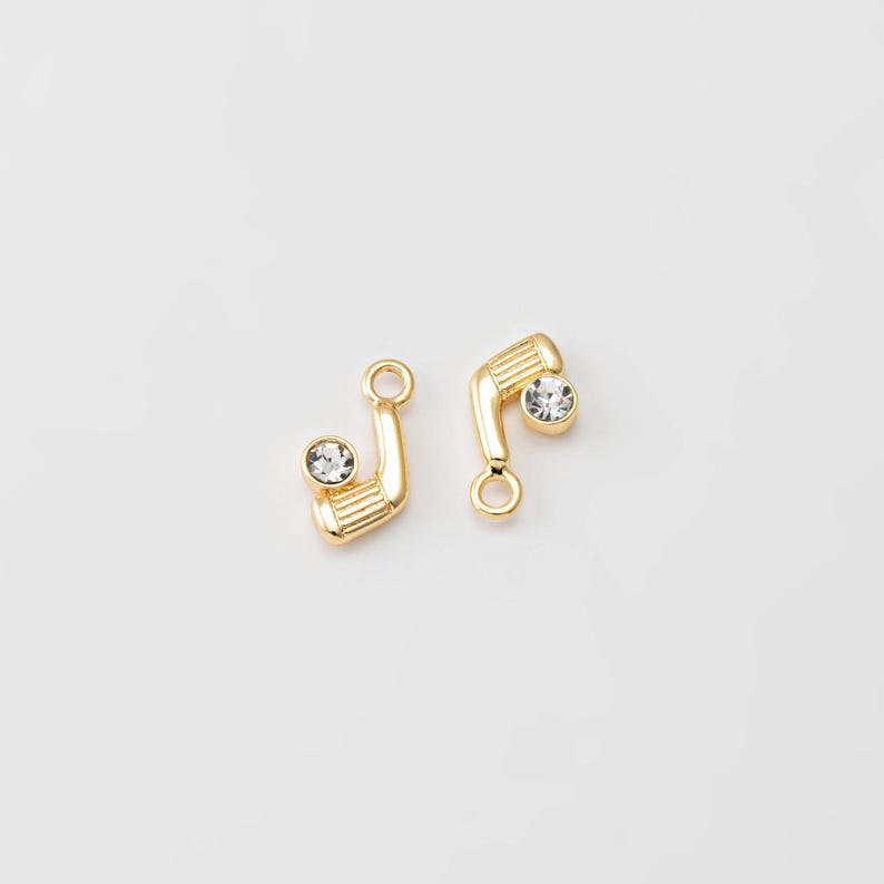 May include: Two gold-colored golf club charms with a clear crystal accent. The charms are detailed with a textured club head and a small loop for attaching to jewelry. The golf club charms are set against a white background.