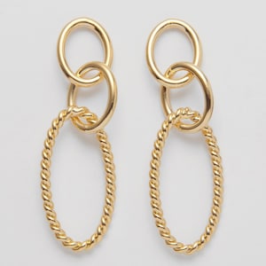 May include: A pair of gold-colored earrings. Each earring features three connected oval shapes. The top two are smooth, while the bottom oval has a twisted rope design. The earrings are displayed against a white background.
