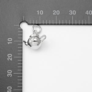 May include: A small, shiny silver teapot charm with a loop for hanging. The teapot has a lid, spout, and handle. The charm is shown against a gray background with measurement markings.