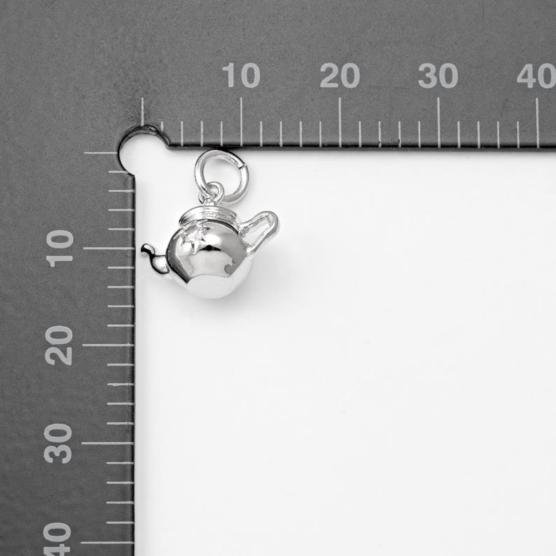 May include: A small, shiny silver teapot charm with a loop for hanging. The teapot has a lid, spout, and handle. The charm is shown against a gray background with measurement markings.