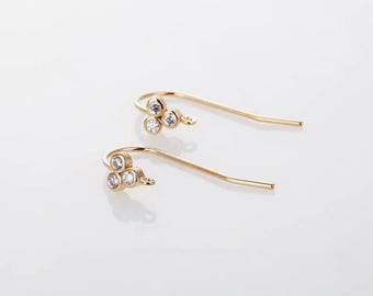 May include: A pair of gold-colored earrings with a hook design. Each earring features three clear, round gemstones clustered together. The earrings are set against a white background, highlighting their delicate design.