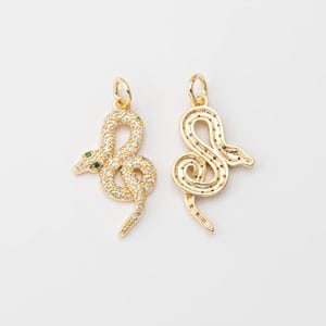 May include: Two gold-colored snake charms. One charm is encrusted with clear and green gemstones, with green gemstones for the eyes. The other charm is a solid gold color with a detailed snake design.