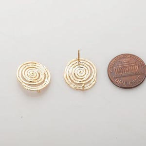 Puede incluir: Dos componentes de pendientes dorados en forma de espiral y una moneda de un centavo estadounidense. Los pendientes tienen una superficie texturizada y un poste para la fijación. La moneda es de color bronce y muestra el Lincoln Memorial.