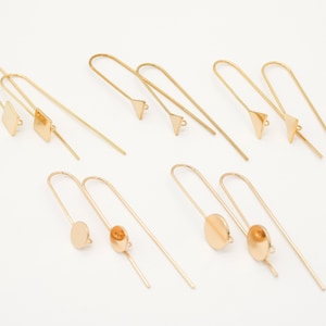 Puede incluir: Un conjunto de componentes de pendientes dorados, con diversas formas geométricas: cuadrados, triángulos y círculos. Los pendientes tienen un diseño de alambre, con un poste para sujetar a la oreja. Los componentes están dispuestos sobre una superficie blanca.