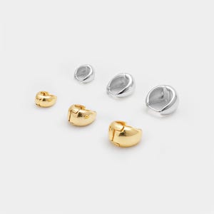 May include: Six earrings in gold and silver are shown on a white backdrop. The earrings are small, chunky hoops with a hinged closure. The gold earrings have a polished finish, while the silver earrings have a shiny, metallic appearance.