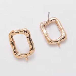 May include: A pair of gold-coloured, rectangular earrings with a textured, organic design. Each earring has a post for piercing and a small loop at the bottom. The earrings are set against a white background.