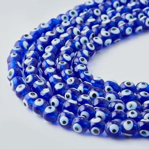 Puede incluir: Primer plano de varios hilos de cuentas de vidrio azul con diseños de ojos blancos y negros. Las cuentas son redondas y tienen un acabado brillante. Las cuentas están dispuestas en un patrón curvo sobre un fondo blanco.
