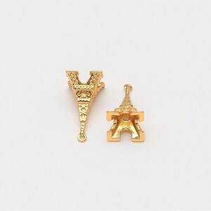 May include: Two gold-coloured Eiffel Tower charms. One charm has a loop at the top and the other has a loop at the bottom.