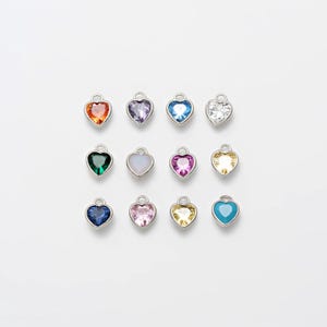 May include: Twelve silver heart-shaped charms with different colored gemstones. The charms are arranged in three rows of four. The colors include orange, purple, blue, clear, green, white, pink, yellow, blue, and turquoise.