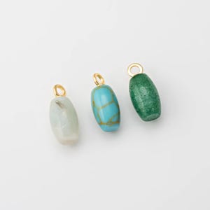 May include: Three gemstone pendants with gold-colored loops. The pendants are oval-shaped and come in three colors: light gray, turquoise with gold veining, and green. The pendants are displayed on a white background.