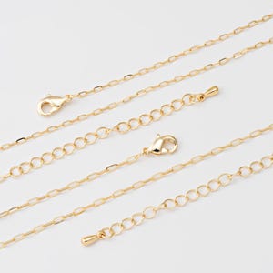 May include: A selection of gold-coloured chain necklaces, featuring lobster claw clasps and extender chains, are shown against a white backdrop. The chains have various link styles, including oval and circular links, suitable for jewellery making.