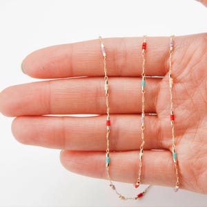 May include: Delicate gold necklaces with colorful beaded accents. The necklaces feature small, rectangular beads in red, turquoise, and lavender, strung along fine gold chains. The necklaces are held in a hand.
