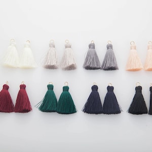 Pode incluir: Uma coleção de borlas coloridas em várias tonalidades, incluindo branco, cinza, pêssego, bordô, verde, azul marinho e preto. Cada borla tem um laço dourado no topo, perfeito para a criação de joias ou projetos de artesanato.