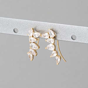 May include: A pair of gold earrings with a teardrop design. Each earring features a row of clear cubic zirconia stones set in a gold setting. The earrings are designed to be worn on the upper ear.