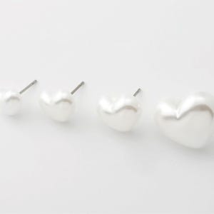 May include: Four white heart-shaped stud earrings of varying sizes. The earrings have a pearlescent finish and are arranged in a row, with the smallest on the left and the largest on the right. The posts are silver-colored.