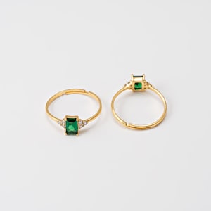 May include: Two gold-colored rings with emerald-cut green gemstones. One ring has a rectangular emerald and small clear stones on each side. The other ring has a similar design. The rings are set against a white background.