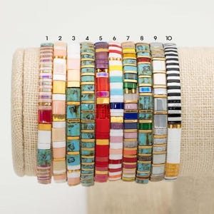 Op de afbeelding: Tien kleurrijke armbanden met verschillende patronen, waaronder turquoise, rood, wit en zwart. De armbanden zijn genummerd van 1 tot en met 10.