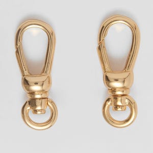 May include: Two gold-coloured swivel clasps. Each clasp features a rounded top with a hinged opening, a cylindrical body, and a circular ring at the bottom. These clasps are designed for attaching to bags, straps, or other accessories.