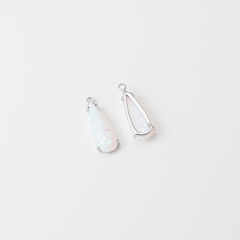 May include: Two teardrop-shaped pendants featuring a white, iridescent stone set in a silver-toned metal frame. Each pendant has a small circular loop at the top for attachment, showcasing a smooth, polished finish and a subtle, shimmering texture.