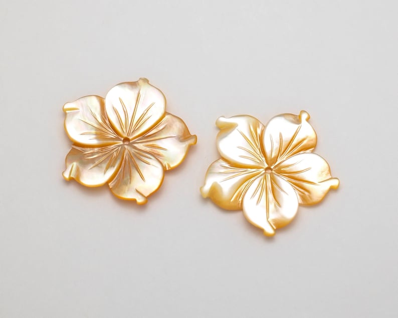 Puede incluir: Dos colgantes iridiscentes en forma de flor. Cada colgante tiene cinco p&eacute;talos con l&iacute;neas grabadas que irradian desde el centro. Las conchas tienen un tono amarillo dorado nacarado, sobre un fondo neutro.