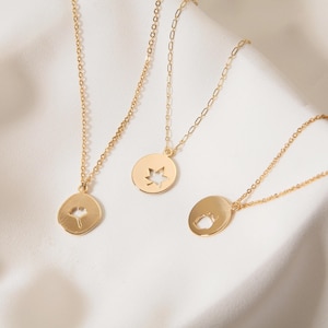 Puede incluir: Tres collares con colgantes dorados con delicadas cadenas. Cada colgante presenta un diseño recortado único: una hoja de ginkgo, una hoja de arce y una pequeña silueta de animal. Los collares se exhiben sobre una superficie blanca texturizada.