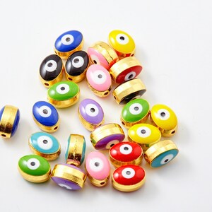 Può includere: Una collezione di perline ovali colorate con una fascia color oro. Le perline presentano un design a occhio centrale in vari colori, tra cui blu, verde, giallo, rosso, rosa, nero e viola. Le perline sono sparse su una superficie bianca.