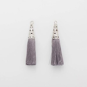 Puede incluir: Dos colgantes de borla grises con tapas cónicas de filigrana plateada. Las borlas están hechas de múltiples hebras de hilo gris. Los colgantes se muestran sobre un fondo blanco.