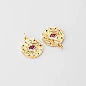 May include: Two gold-toned pendants with a circular design featuring a stylized eye with a red gemstone surrounded by smaller colorful gemstones.