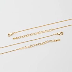 Puede incluir: Tres collares dorados con delicadas cadenas y cadenas extensoras. Cada collar presenta un cierre de mosquetón para una sujeción segura. Los collares se muestran sobre un fondo blanco liso, resaltando su simple elegancia.