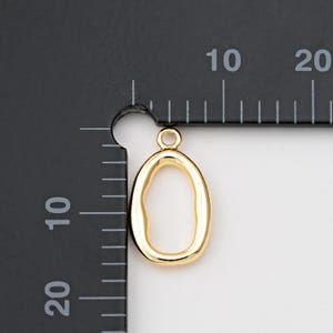 Puede incluir: Un colgante dorado con forma ovalada irregular y un peque&ntilde;o lazo en la parte superior. El colgante se muestra junto a una regla con marcas en mil&iacute;metros, lo que sugiere su tama&ntilde;o. El colgante es probablemente para la fabricaci&oacute;n de joyas o manualidades.