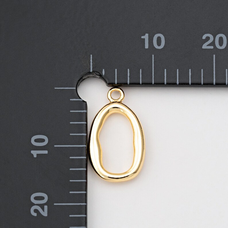 Puede incluir: Un colgante dorado con forma ovalada irregular y un peque&ntilde;o lazo en la parte superior. El colgante se muestra junto a una regla con marcas en mil&iacute;metros, lo que sugiere su tama&ntilde;o. El colgante es probablemente para la fabricaci&oacute;n de joyas o manualidades.