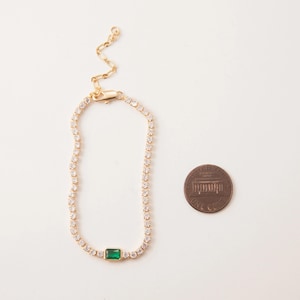 May include: A gold-toned bracelet with a row of small, clear crystals and a rectangular emerald-green stone in the center. A penny is next to the bracelet for size comparison. The bracelet has an adjustable chain.