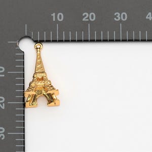 May include: Gold-coloured metal charm shaped like the Eiffel Tower. The charm has a loop at the top for attaching to a necklace or bracelet.